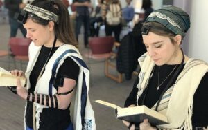 two Jewish women, one trans, praying in public wearing tefilin. They're also wearing prayer shawls and holding siddurs.